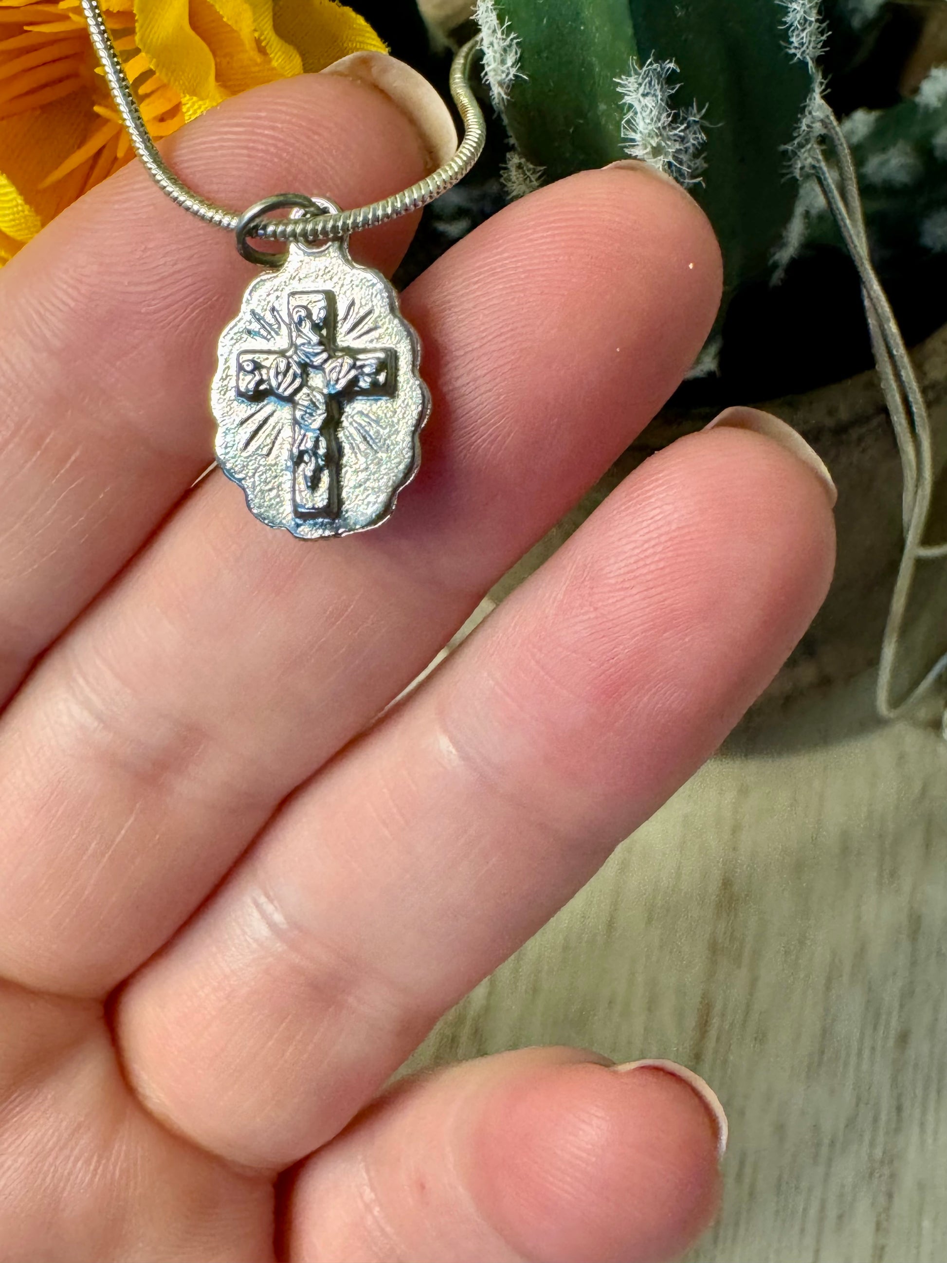 Silver pendant with a religious design held between fingers, with a blurred background of yellow flowers and greenery.